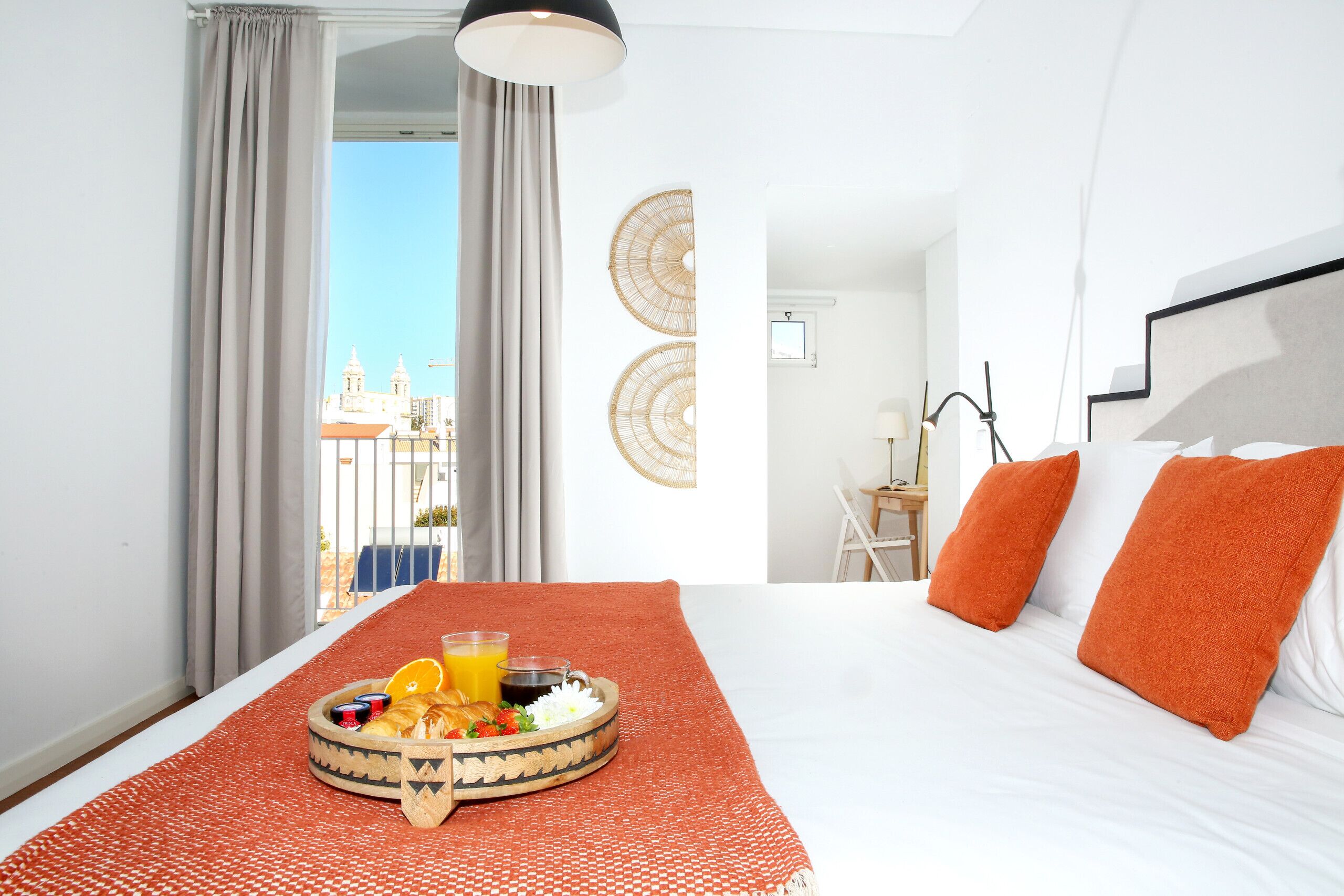 A hotel room with a bed, orange pillows, a breakfast tray with orange juice, and a view of buildings outside.