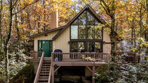 A cabin with a green door, a deck, and a staircase leading up to it, surrounded by trees. - COZY A-Frame Spacious Deck w/ Grill (Wintergreen)