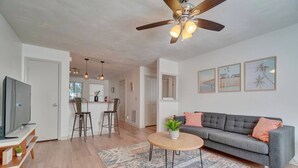 A modern living room with a grey sofa, a coffee table, and a TV on a stand. There is a ceiling fan, a kitchen area with bar stools, and a patterned rug. - King Bed WIFI - Work Space - 2 parking space B (Norfolk)