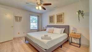 A bedroom with a bed, a ceiling fan, a bedside table with a lamp, a hanging plant, and a window with curtains. - King Bed WIFI - Work Space - 2 parking space B (Norfolk)