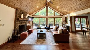 A spacious living room with a stone fireplace, a large ceiling fan, and a view of the outdoors through large windows.