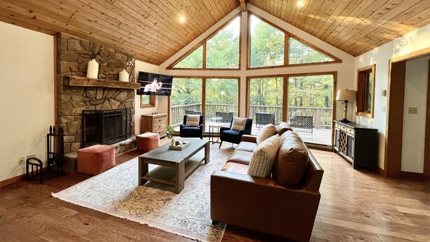 A spacious living room with a stone fireplace, large windows, and a wooden ceiling.