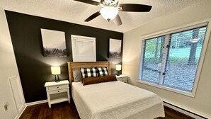 A bedroom with a bed, two bedside tables, a ceiling fan, and a large window with blinds.