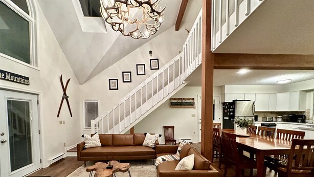 A spacious living area with a brown sectional sofa, a wooden coffee table, and a dining table with chairs. A staircase leads to an upper level.