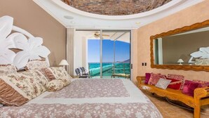 Quarto com uma cama grande, um banco de madeira e uma varanda com vista para o oceano.