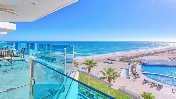 Varanda com guarda-corpo de vidro, vista para uma praia e uma piscina.