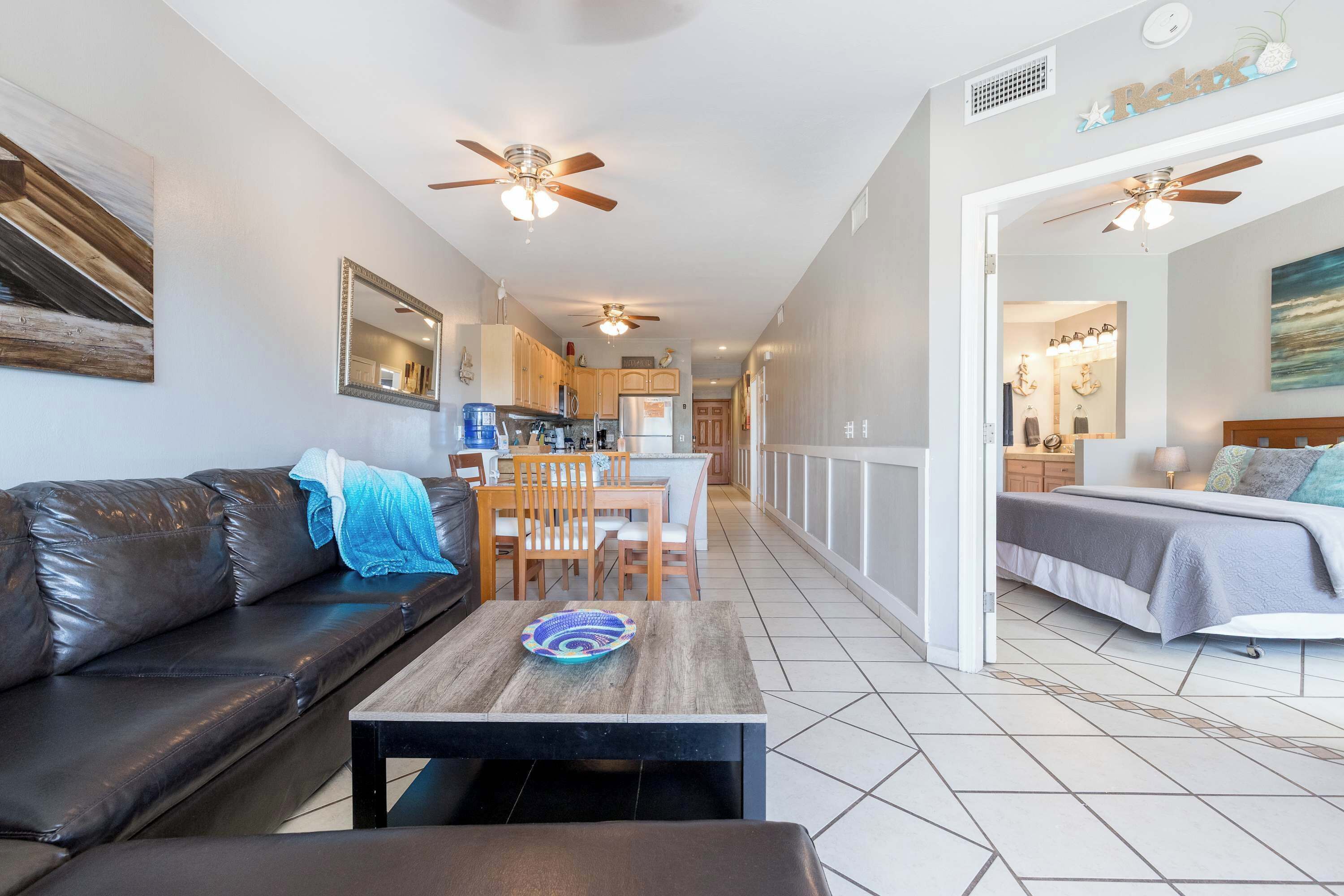A modern living room with a leather sofa, a wooden coffee table, and a dining area with wooden chairs. There is a kitchen in the background with a refrigerator and cabinets.