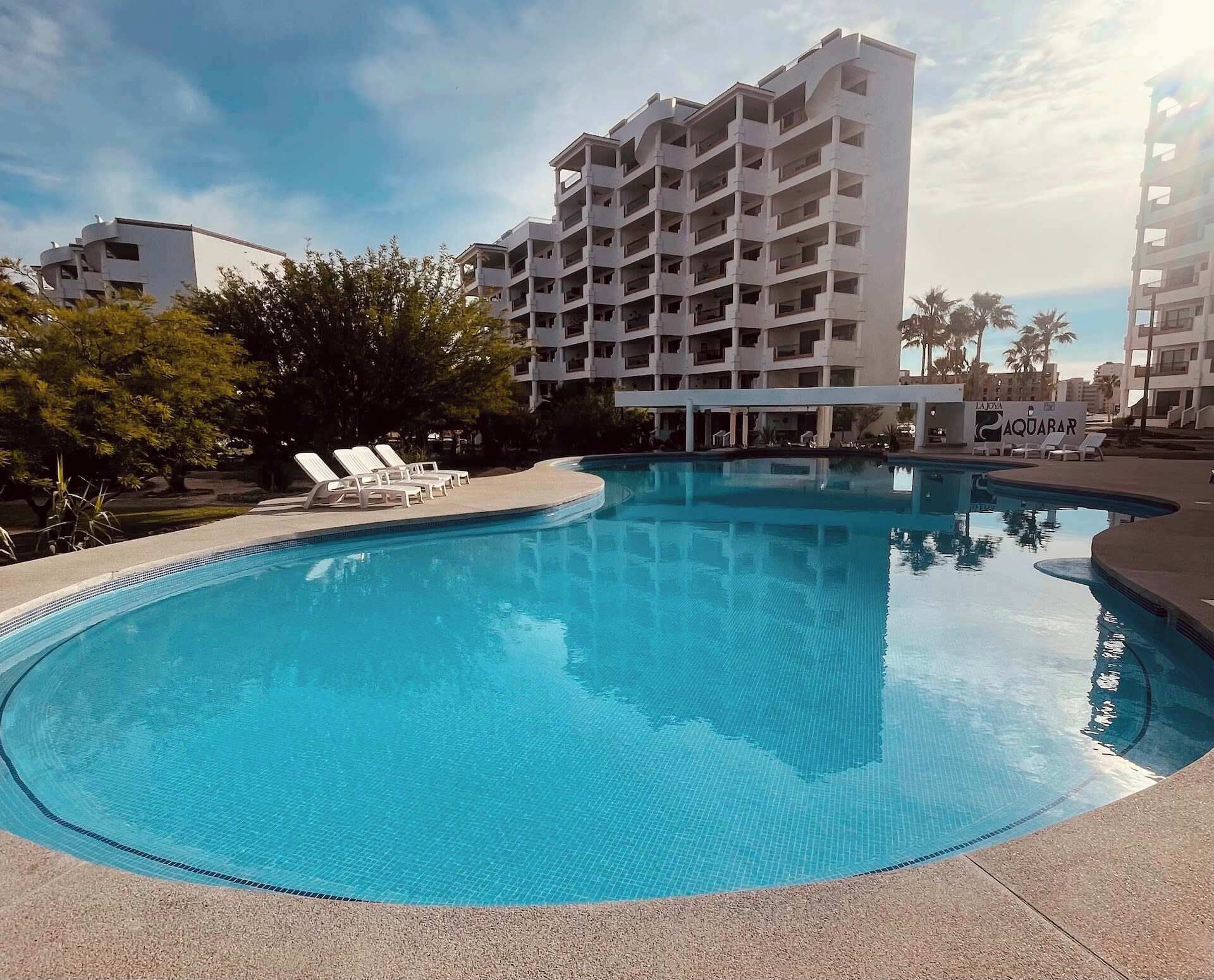 Une piscine entourée de chaises longues, elle-même bordée d’un bâtiment doté de nombreux balcons et de palmiers.