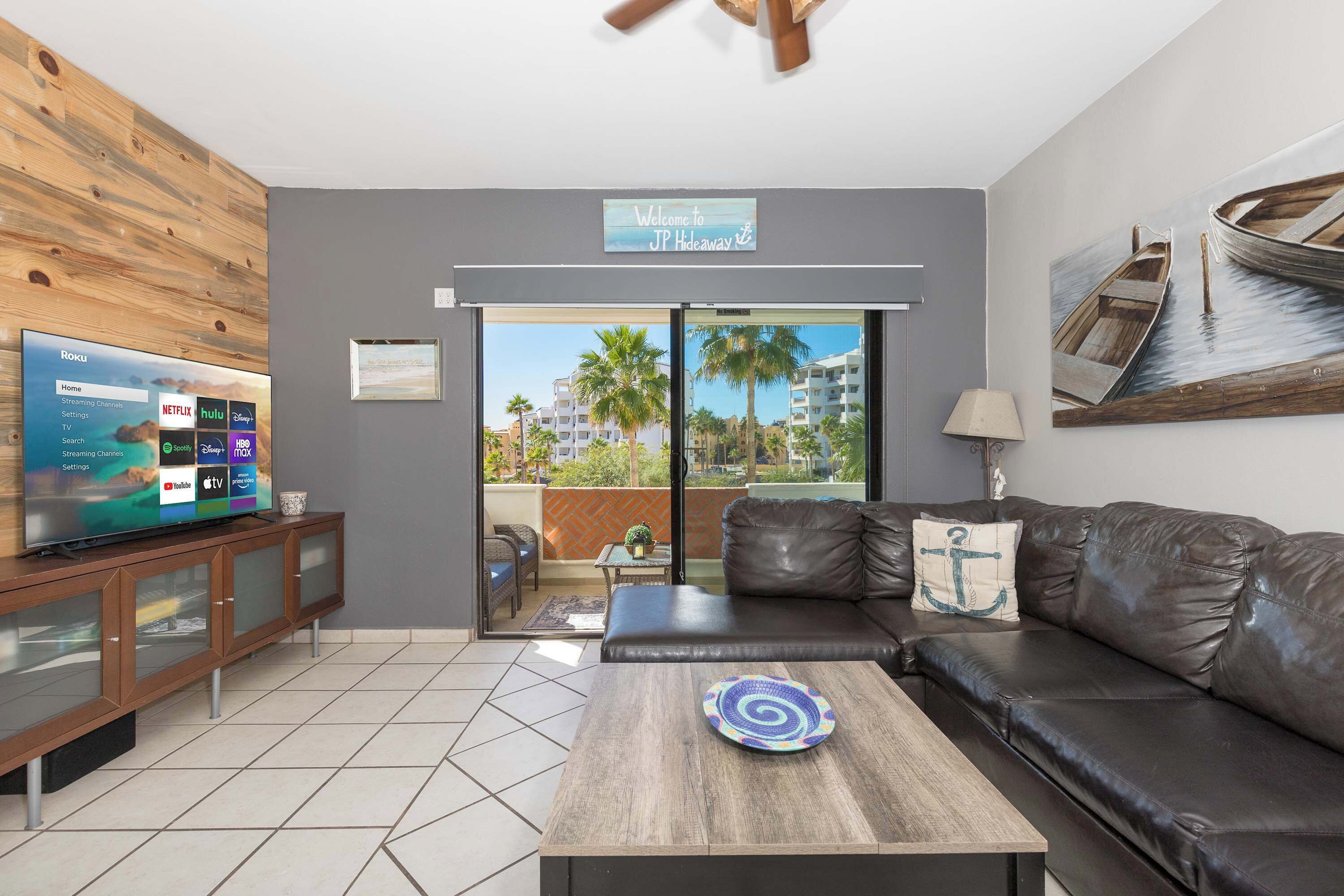 A modern living room with a flat-screen TV displaying streaming services, a leather sofa, a coffee table, and a balcony with a view of palm trees and buildings.