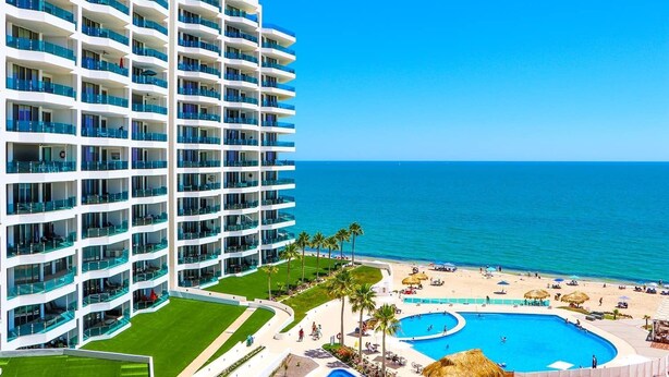 A high-rise hotel with multiple balconies overlooking a beach and a pool area.