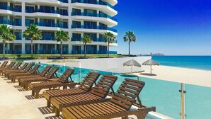 A resort pool area with wooden lounge chairs and a clear view of the beach and ocean.