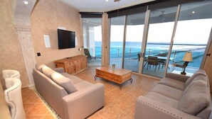 A spacious living room with two sofas, a coffee table, and a TV mounted on the wall. The room has a large sliding glass door leading to a balcony with a view of the ocean.
