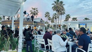 Zona pranzo all'aperto con sedie e tavoli bianchi, circondata da palme e un edificio con un cartello che riporta la scritta "Joya Bistro & Bar".