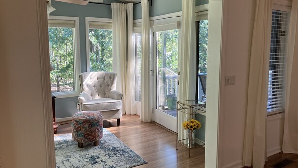 Ein Zimmer mit einem großen Fenster, einem weißen Sessel, einem gemusterten Hocker und einem Metall-Pflanzentisch mit gelben Blumen.