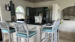 Een keuken met een centraal eiland, donkere kastjes en lichtgekleurde stoelen met turquoise kussens.