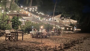 Een buitenterras met tafels en parasols, omringd door bomen en lichtjes.