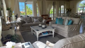 Een ruime woonkamer met een grote witte bank, een salontafel en een eethoek met rieten stoelen.