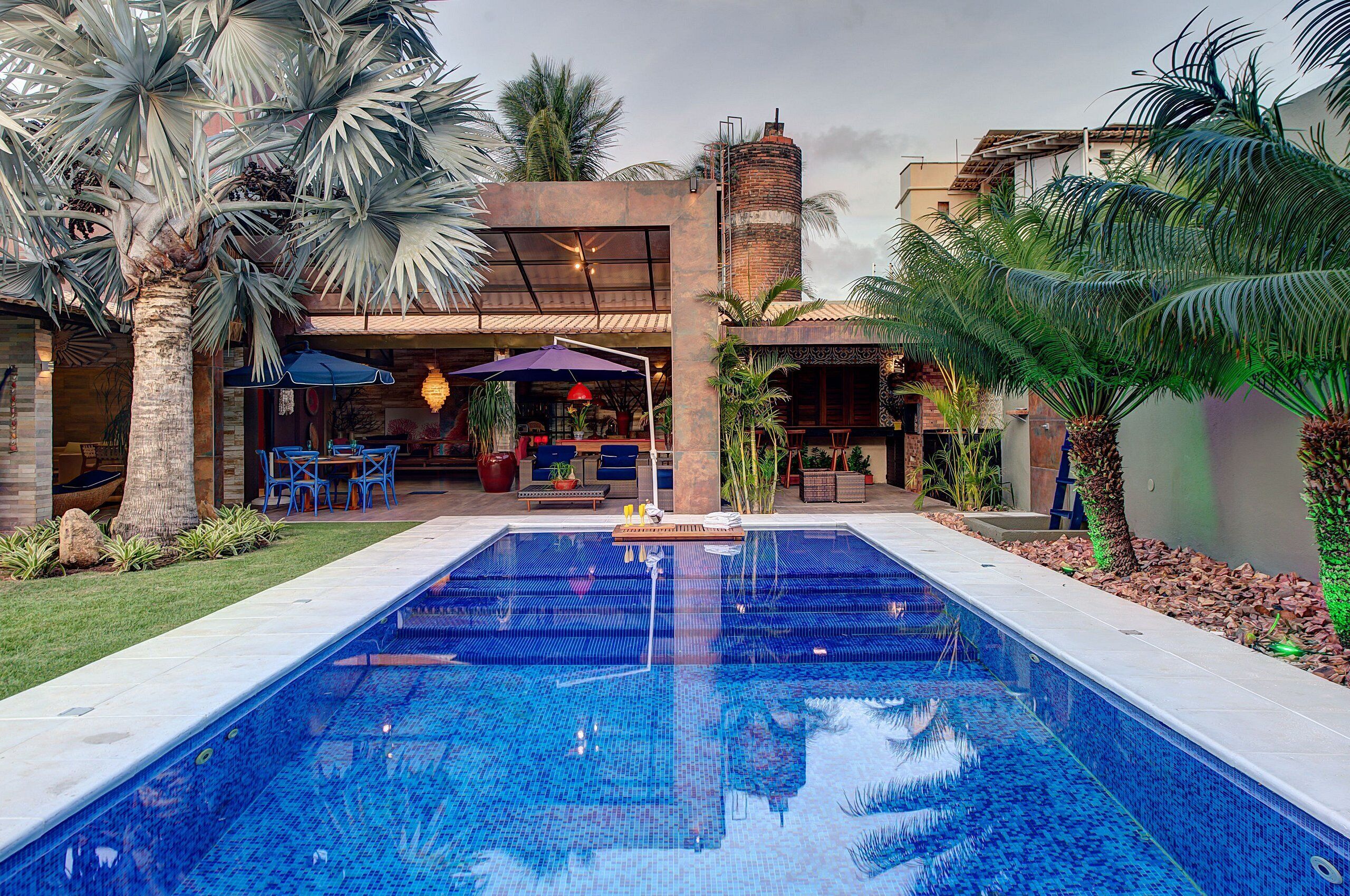 A swimming pool with a tiled deck, surrounded by palm trees and a building with outdoor seating.