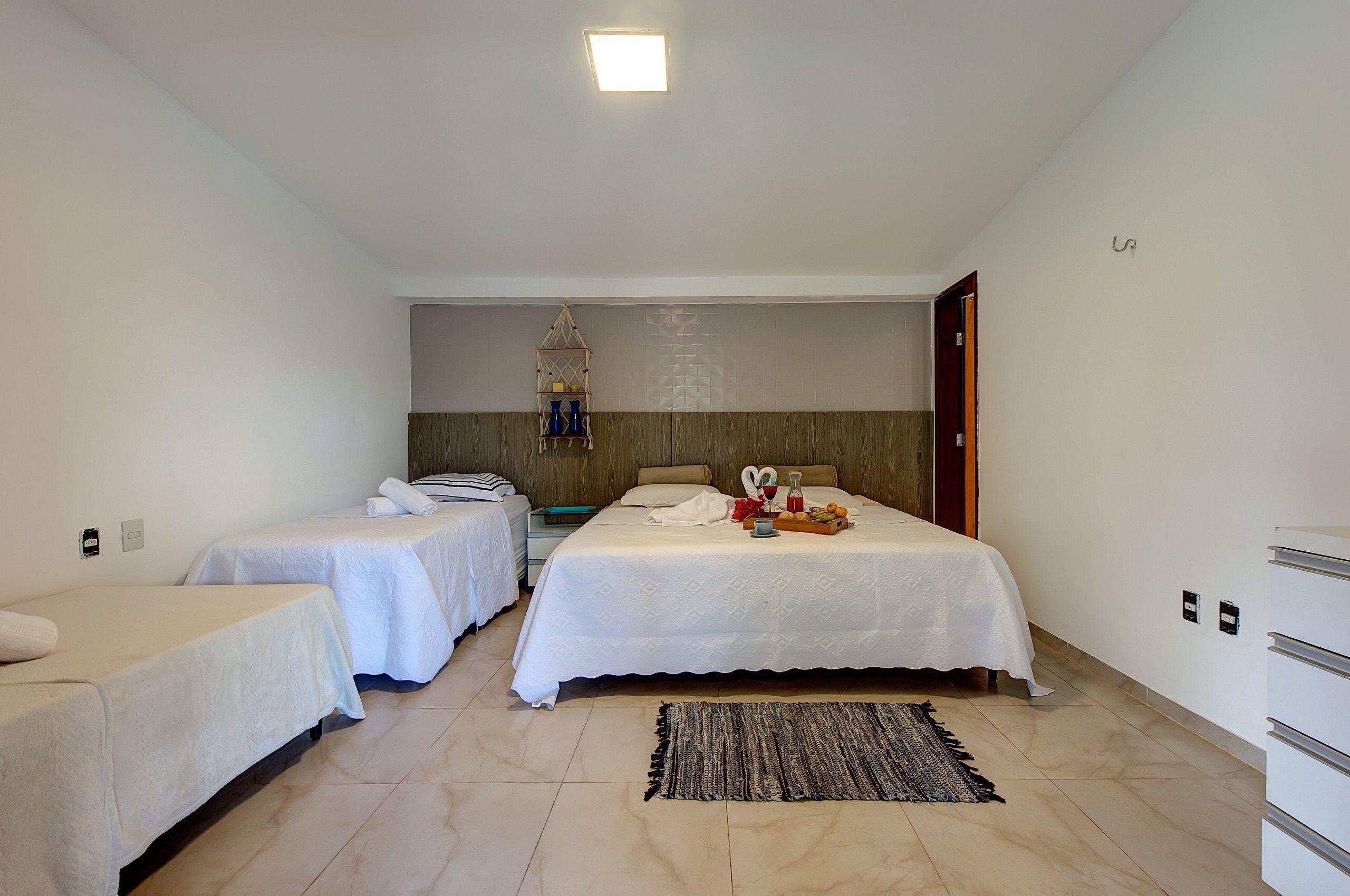 A hotel room with two single beds, a large bed, a bedside table with a lamp, a wall-mounted shelf, and a door to the right.