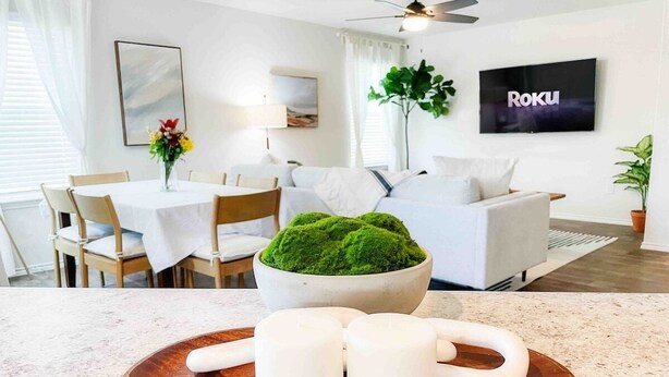 Una sala moderna con un sofá blanco, una mesa de comedor con sillas, un televisor montado en la pared y un recipiente con musgo verde en el suelo.