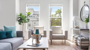 Uma sala de estar moderna com um sofá cinzento, duas poltronas, uma mesa de centro com uma tigela e revista, e vista para um edifício através de grandes janelas.