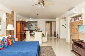 Amplia sala de estar con una cama grande, ventilador de techo, televisor y una cocina al fondo.