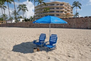 A beach with two blue lounge chairs under a blue umbrella, a stone wall, and a multi-story building in the background. - Ocean Front Condo with Pool Sleeps 6 People (Nuevo Vallarta)