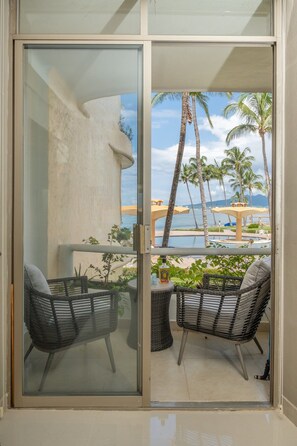 A balcony with two wicker chairs, a small table with a bottle, and a view of a beach with palm trees and a pool. - Ocean Front Condo with Pool Sleeps 6 People (Nuevo Vallarta)
