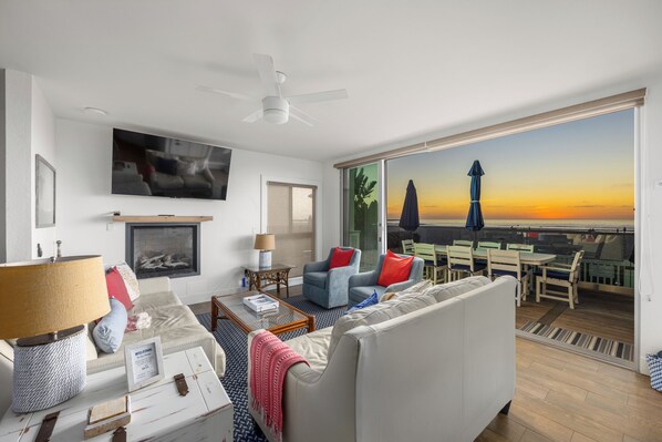 Un salon moderne avec un canapé, des fauteuils, une table basse et un téléviseur. Il y a un balcon avec vue sur le coucher du soleil, ainsi qu'une table et des chaises à l'extérieur.