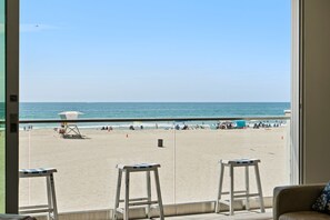 Uitzicht op een strand vanaf een kamer met een groot raam, met een zandstrand, heldere lucht en enkele strandstoelen en parasols.