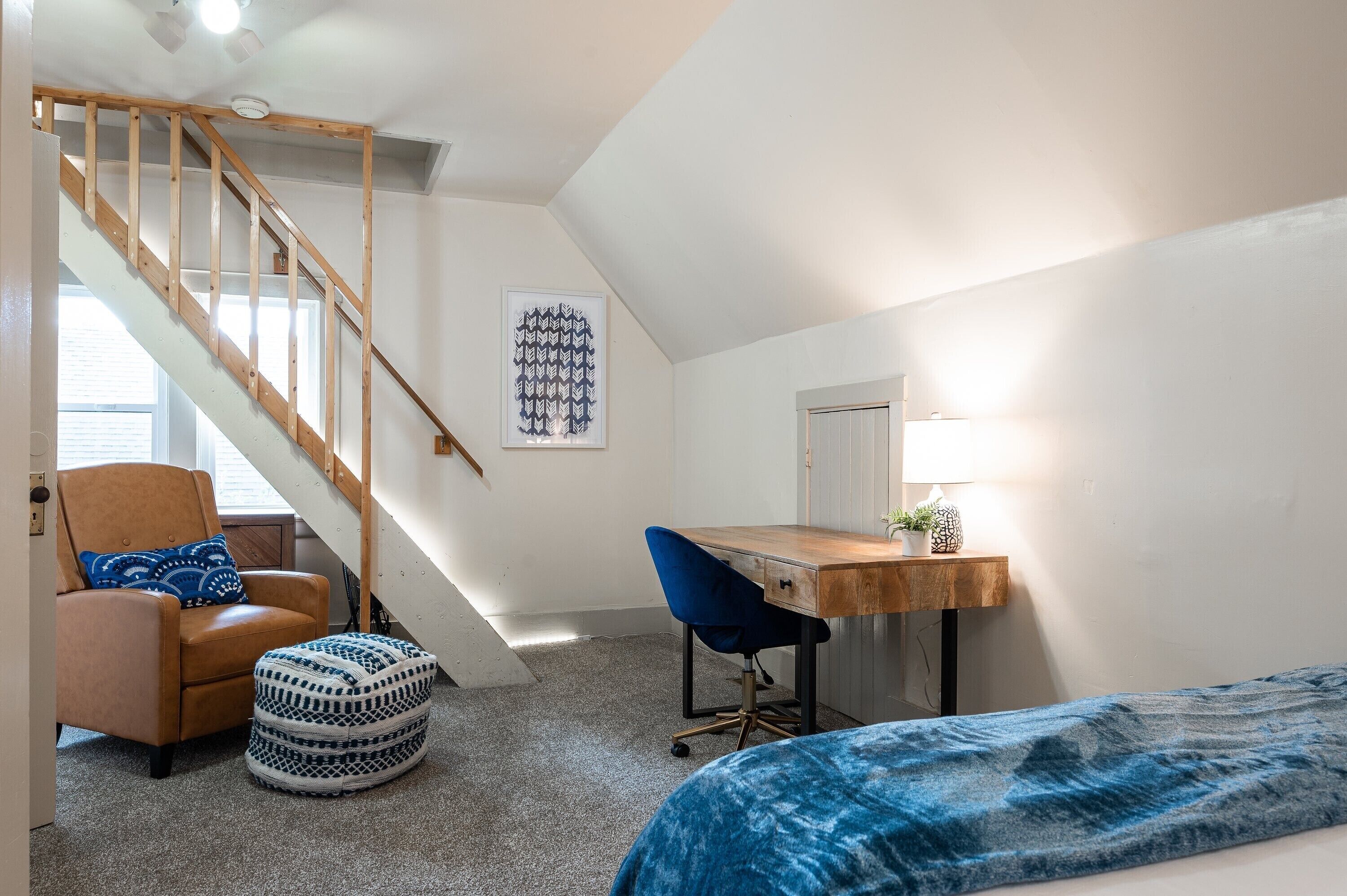 A bedroom with a wooden staircase, a desk, a chair, a bed, and a patterned ottoman.