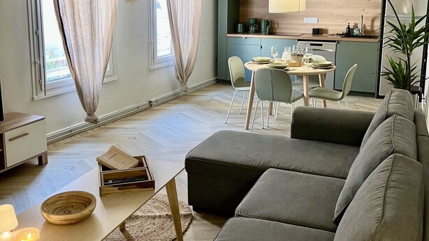 Moderni olohuone, jossa on harmaa kulmasohva, pieni ruokapöytä ja taustalla keittiötila.