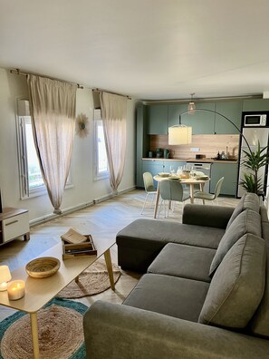 Ein modernes Wohnzimmer mit einem grauen Ecksofa, einem kleinen Esstisch und einer Küchenzeile im Hintergrund.