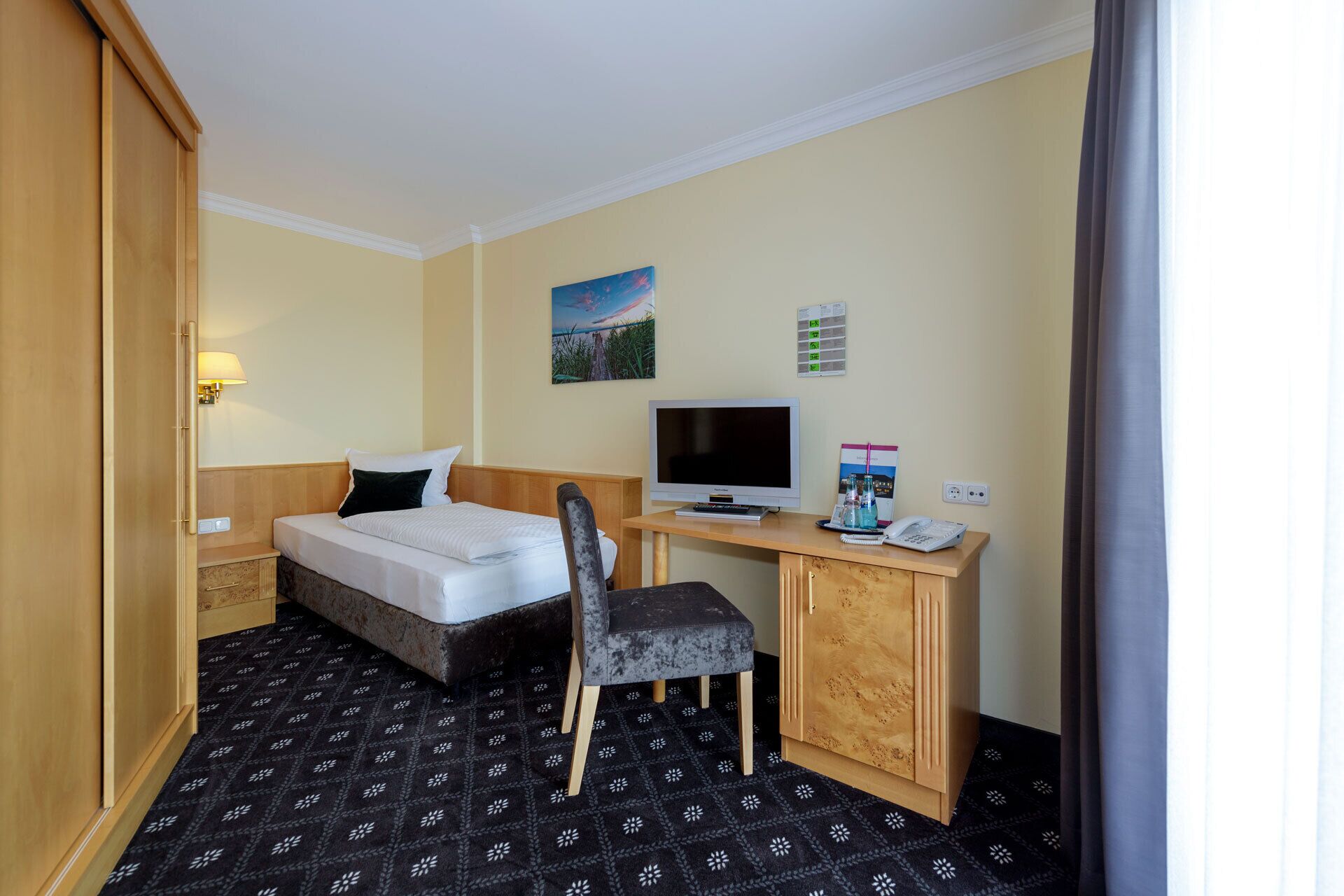 A hotel room with a bed, a desk with a computer, a chair, a TV, and a wall-mounted phone.