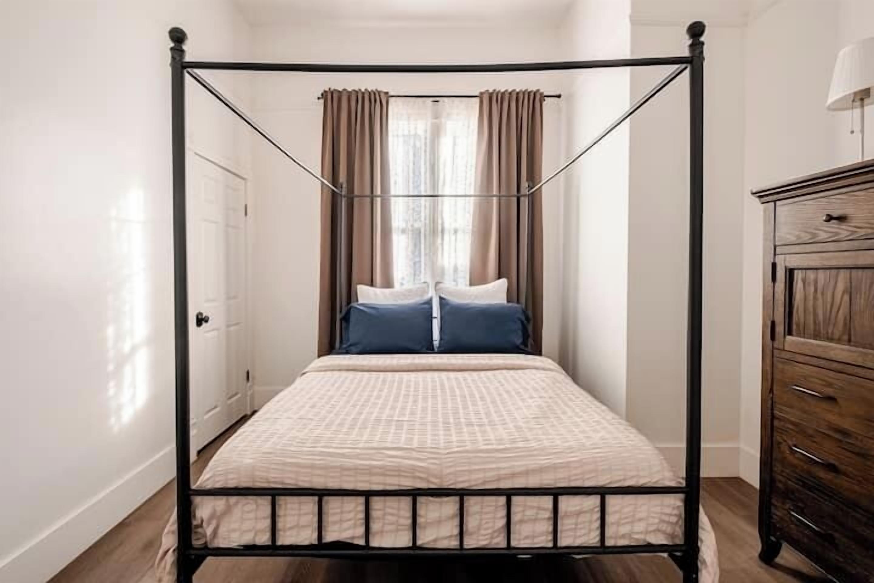 A four-poster bed with a beige bedspread, two pillows, and a wooden dresser to the right.
