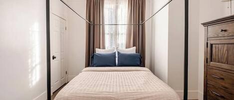 A four-poster bed with a beige bedspread, two pillows, and a wooden dresser to the right.