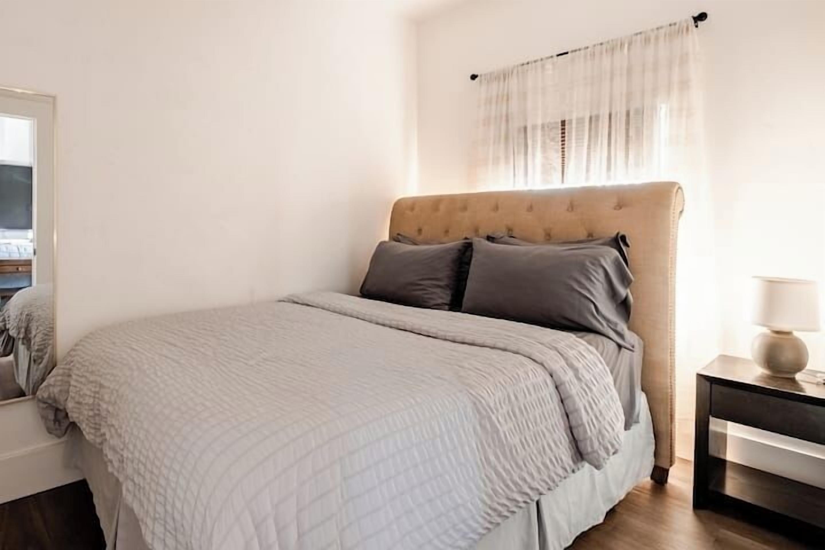 A bedroom with a bed, a nightstand, and a window with sheer curtains.