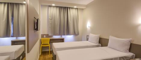 A hotel room with two single beds, a small desk, a chair, and a window with curtains.