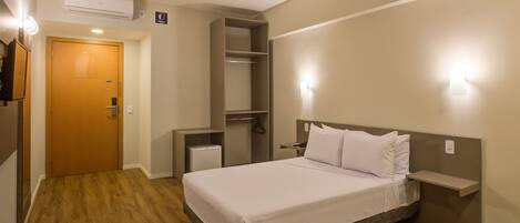 A hotel room with a bed, a wardrobe, a bedside table, and a wall-mounted shelf.