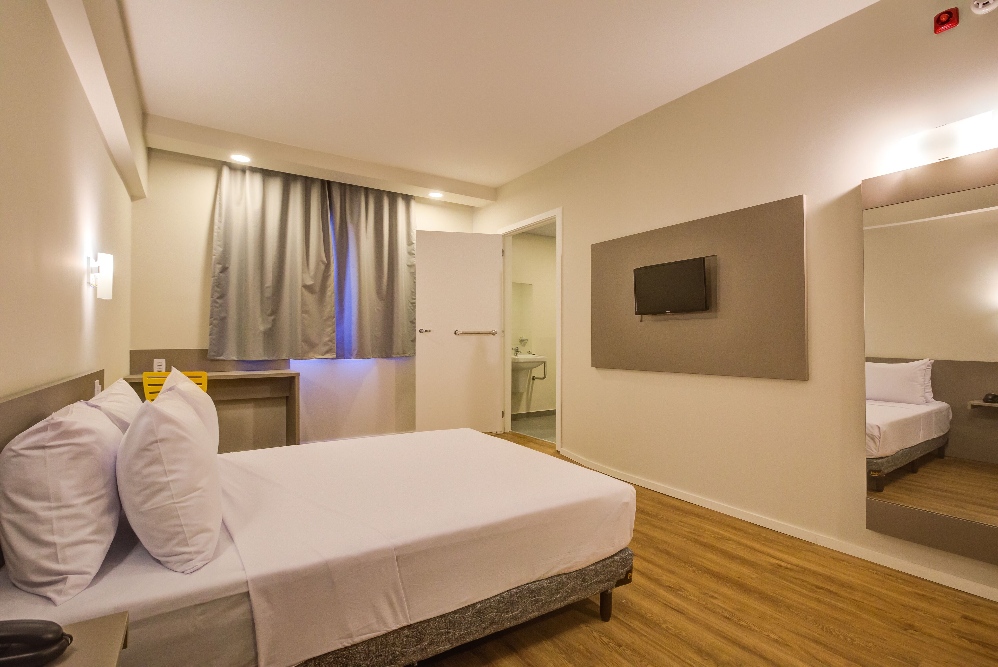 A hotel room with a bed, a bedside table, a mirror, a television mounted on the wall, and a bathroom visible through an open door.