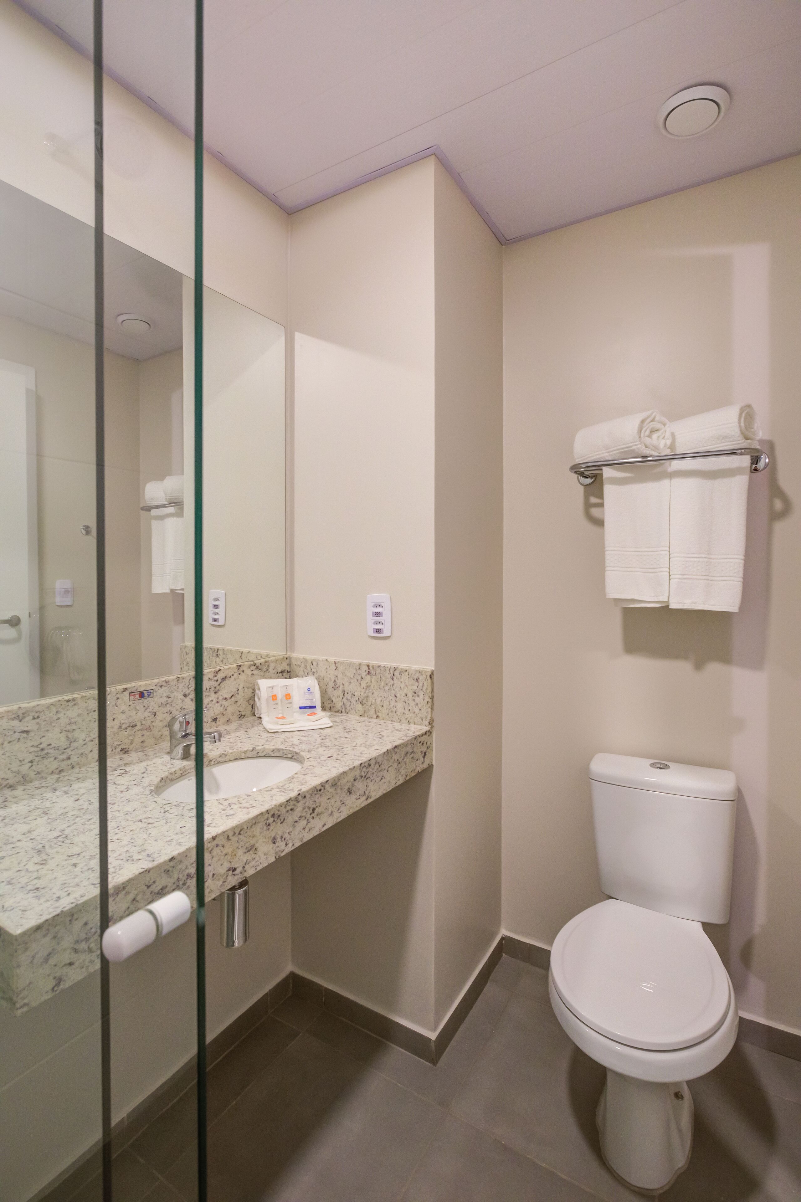 Un baño moderno con cabina de ducha de vidrio, lavamanos de mármol y un inodoro con toallero encima.
