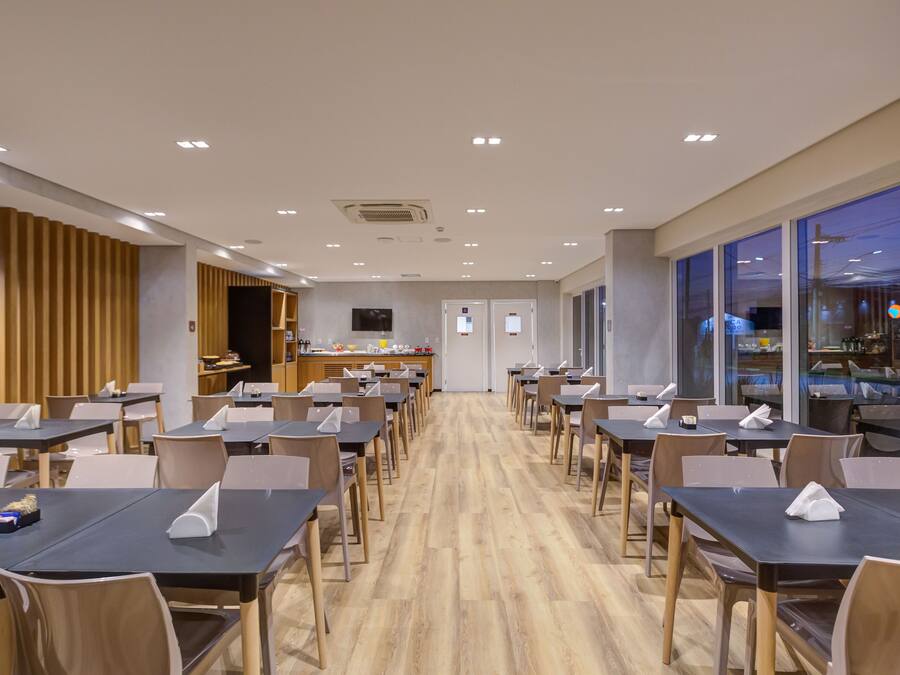 A modern dining area with multiple tables and chairs, a kitchen in the background, and large windows on one side.