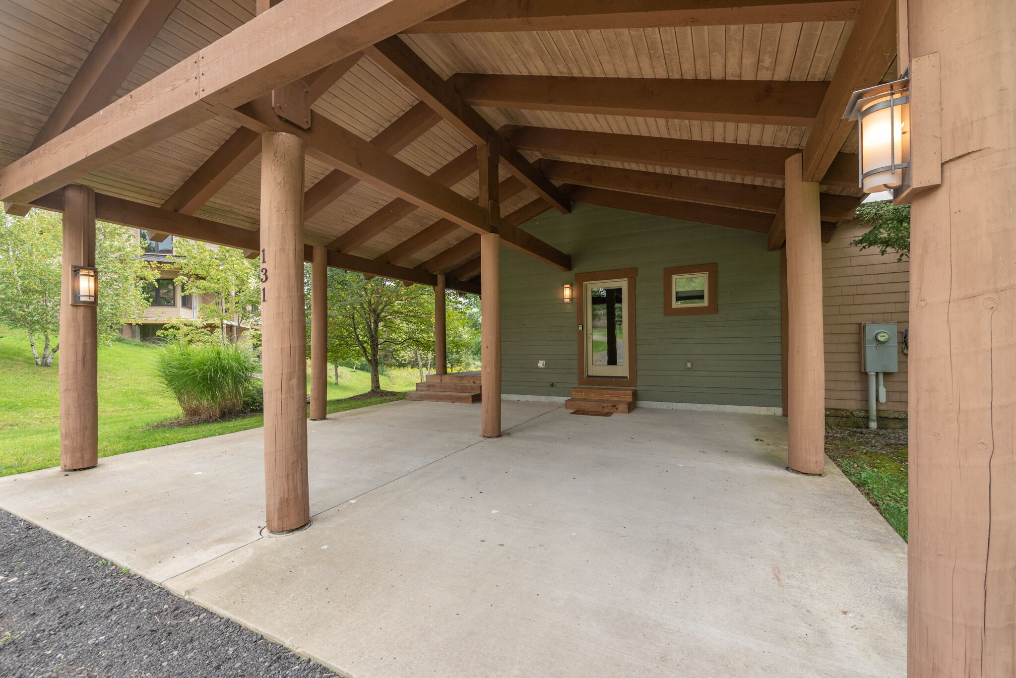 A covered porch with wooden beams and a concrete floor, leading to a green building with a door and windows.