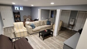 Una sala de estar con sofá, sillón y una mesa de centro. Se observa una puerta, una estantería y una ventana.