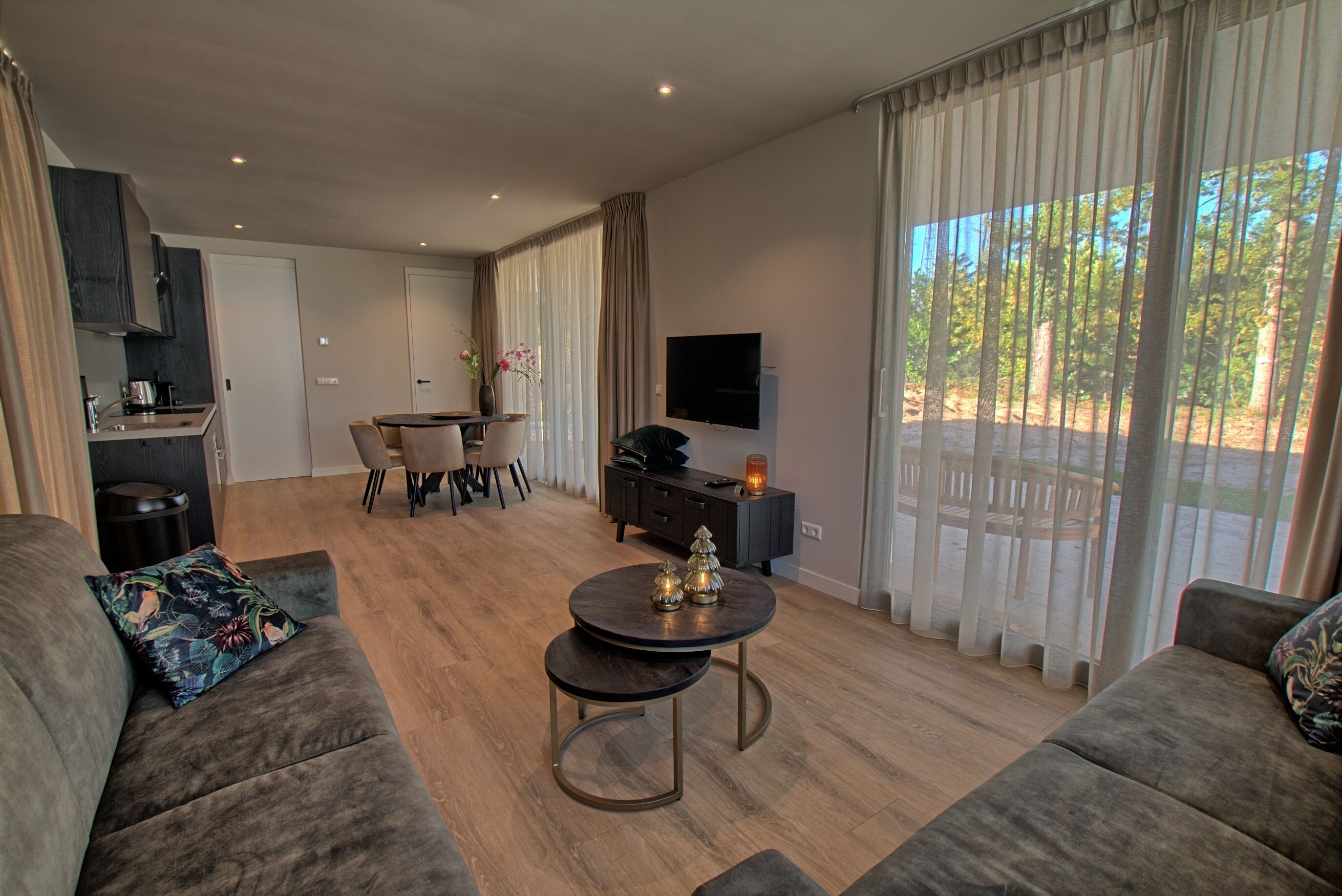 Una sala de estar moderna con sofá, una mesa redonda de centro y un comedor con mesa y sillas. Se observa una cocina con armarios y un televisor montado en la pared.