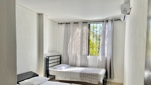 A bedroom with two beds, a window with curtains, a bedside table, and a wall-mounted air conditioning unit. - Beautiful and cozy apartment in Mompós (Santa Cruz de Mompox)