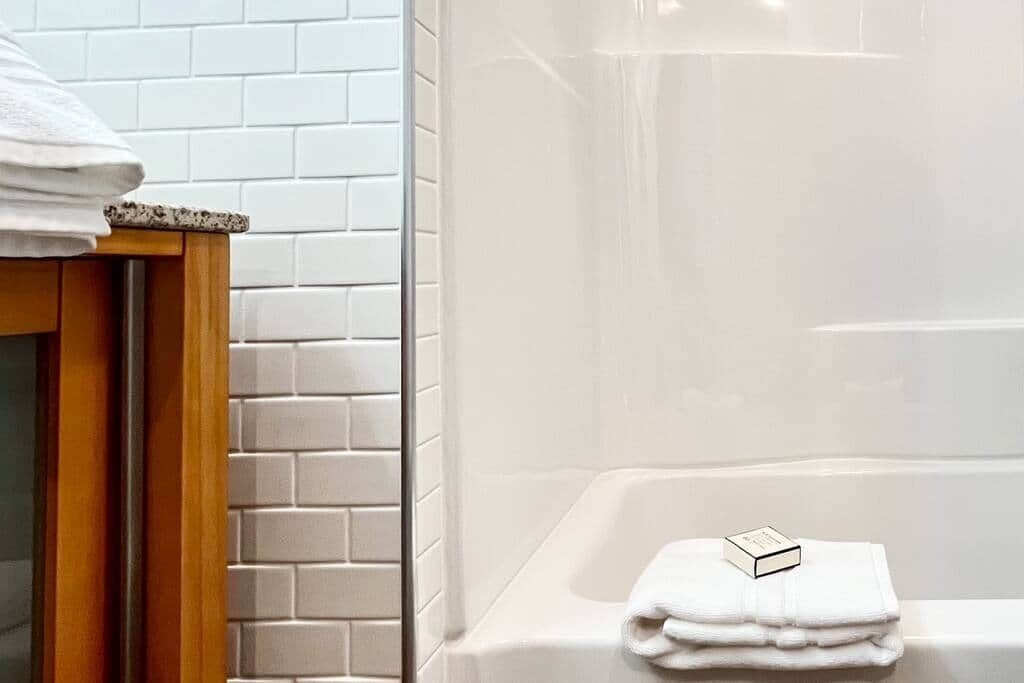 A bathroom with white tiled walls, a wooden cabinet, and a white bathtub with a folded towel and a soap bar on it.