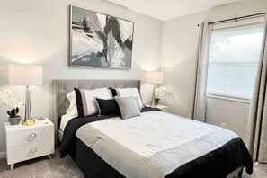 A bedroom with a bed, two bedside tables, a nightstand, a wall-mounted painting, and a window with curtains. - Sunsets on the Grand (Lansing)