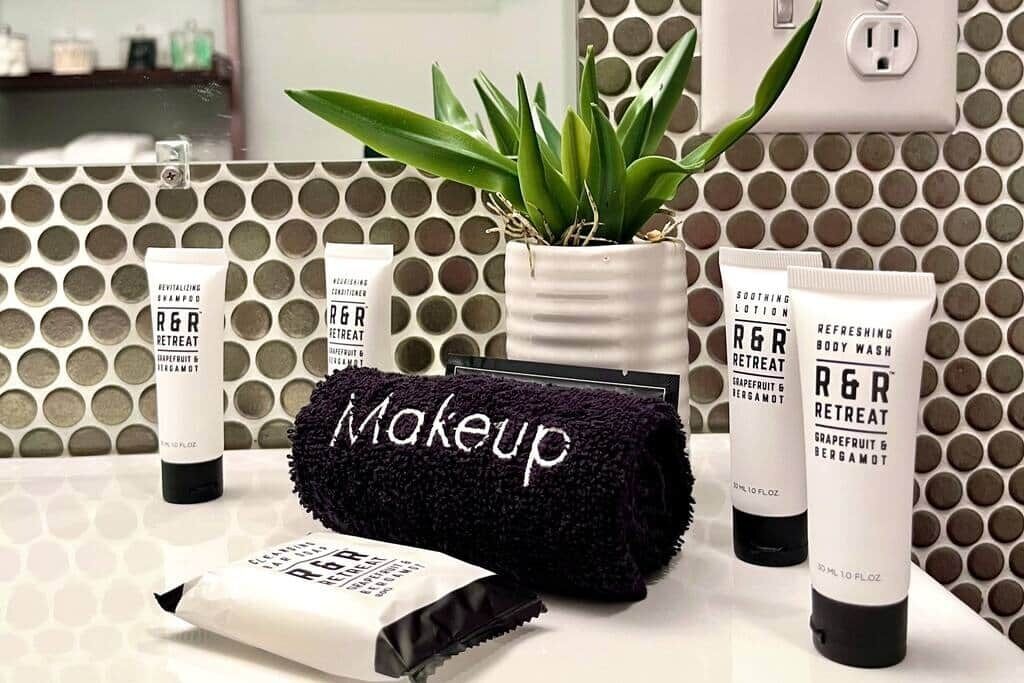 A bathroom with a patterned tile wall, a potted plant, and various R&R brand toiletries.
