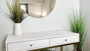 A white console table with two drawers, a round mirror above, and two potted plants on either side. - Sunsets on the Grand (Lansing)